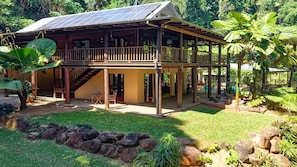 A two-story wooden house with a balcony, solar panels, and a garden with rocks and plants. - Fern House your serene rainforest retreat (Bingil Bay)