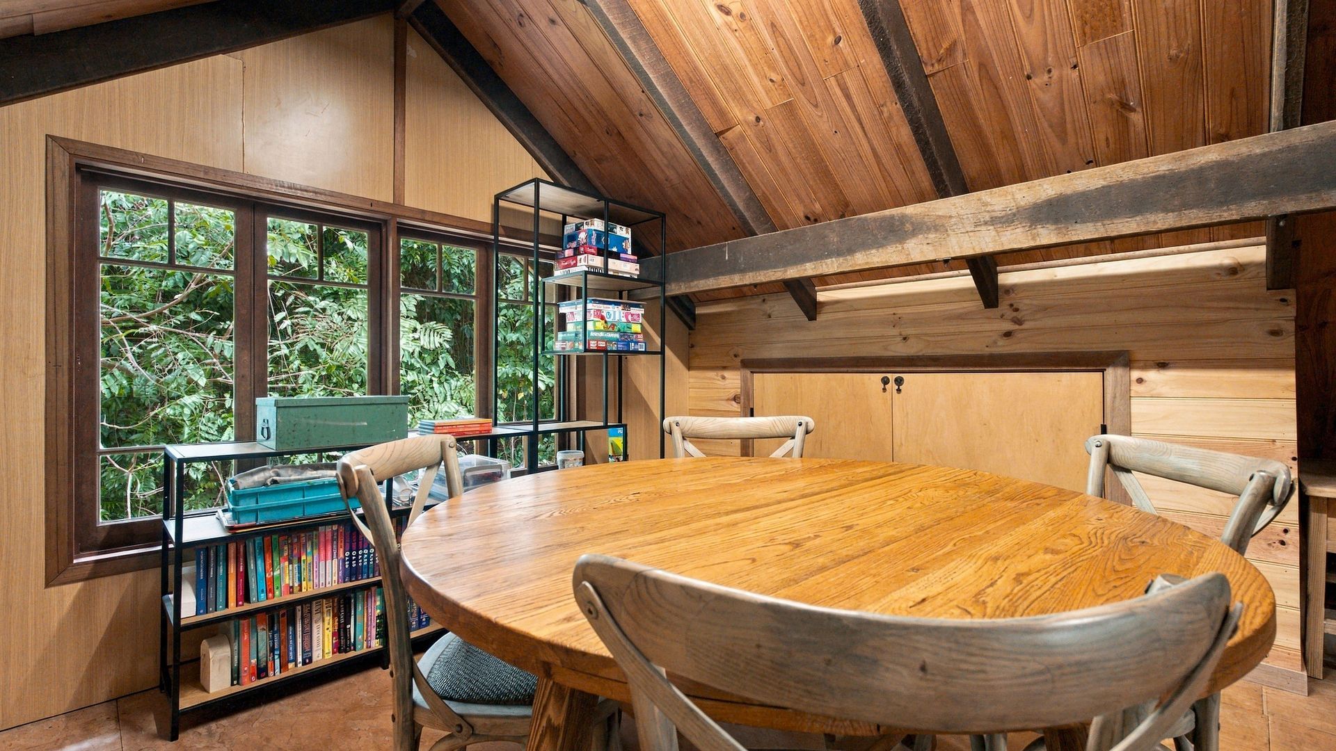 A wooden interior with a round table and chairs, bookshelves, and a view through large windows.