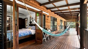 A wooden deck with a hammock, a bed with patterned bedding, and a view of the outdoors.