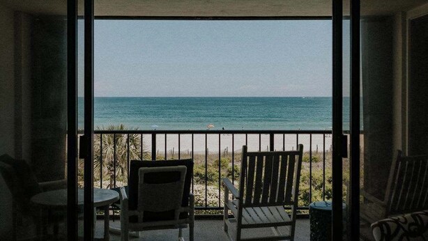A balcony with a view of the ocean, featuring two wooden chairs and a small table. - The Spoleto by Mira-Mar (Wrightsville Beach)