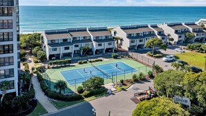 A tennis court surrounded by apartment buildings with a view of the ocean. - The Spoleto by Mira-Mar (Wrightsville Beach)