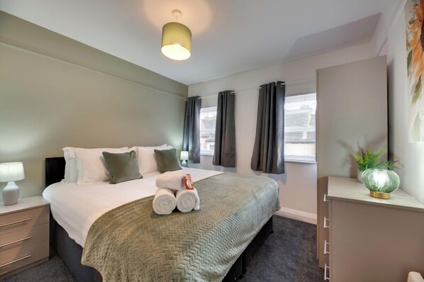 Une chambre d’hôtel avec un lit, une table de chevet, une commode, une lampe et une fenêtre avec des rideaux.