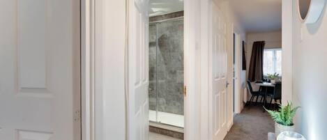 A hallway with a glass shower, a dining area with a table and chairs, and a small plant on a console table.