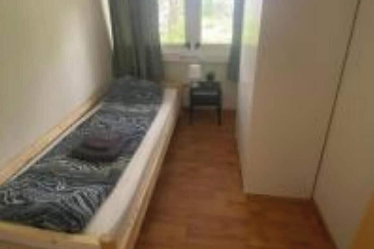A small, single-bed room with a wooden floor, a window with curtains, and a small table.
