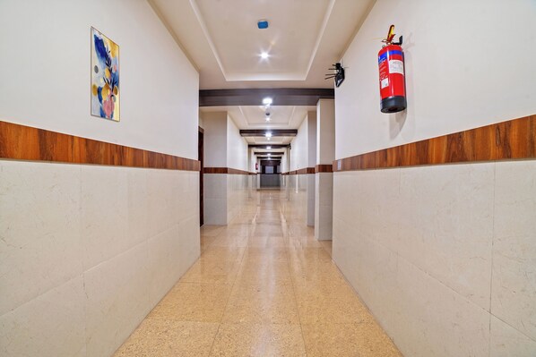 Un pasillo largo y bien iluminado, con un extintor de incendios montado en la pared, un panel de madera en la parte inferior y una obra enmarcada a la izquierda.