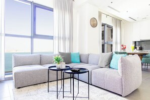 Una sala de estar moderna con un sofá seccional gris, dos mesas redondas negras y vistas al mar a través de amplios ventanales.