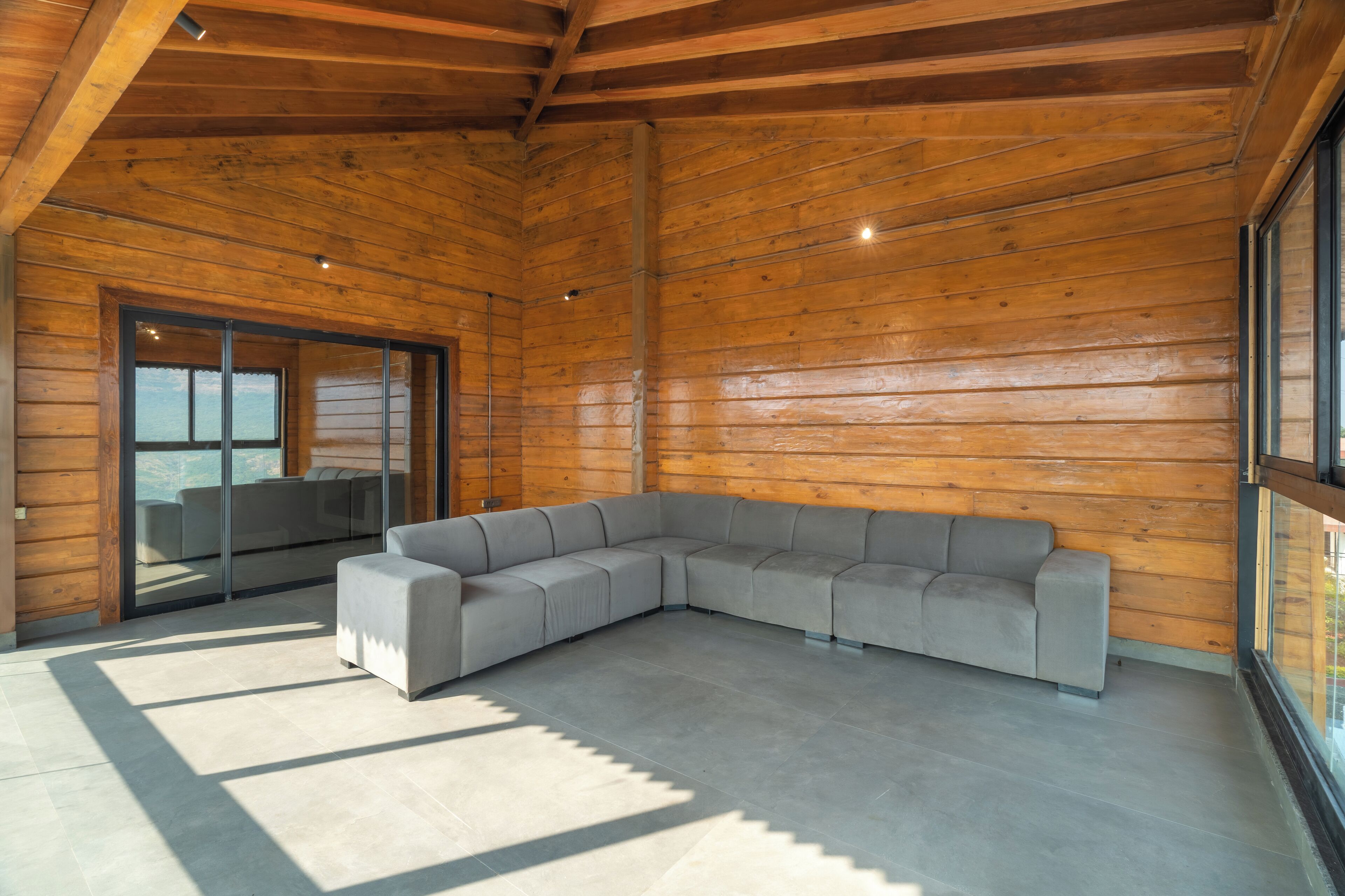 A modern log cabin interior with a large sectional sofa, a sliding glass door leading to a balcony, and a high ceiling with exposed wooden beams.