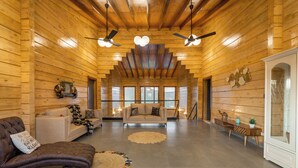 A spacious log cabin interior with a high ceiling, two ceiling fans, a large window, a sofa, a loveseat, and a wooden cabinet. - Villa 199 Amarja's Sunset Retreat (Mawal)