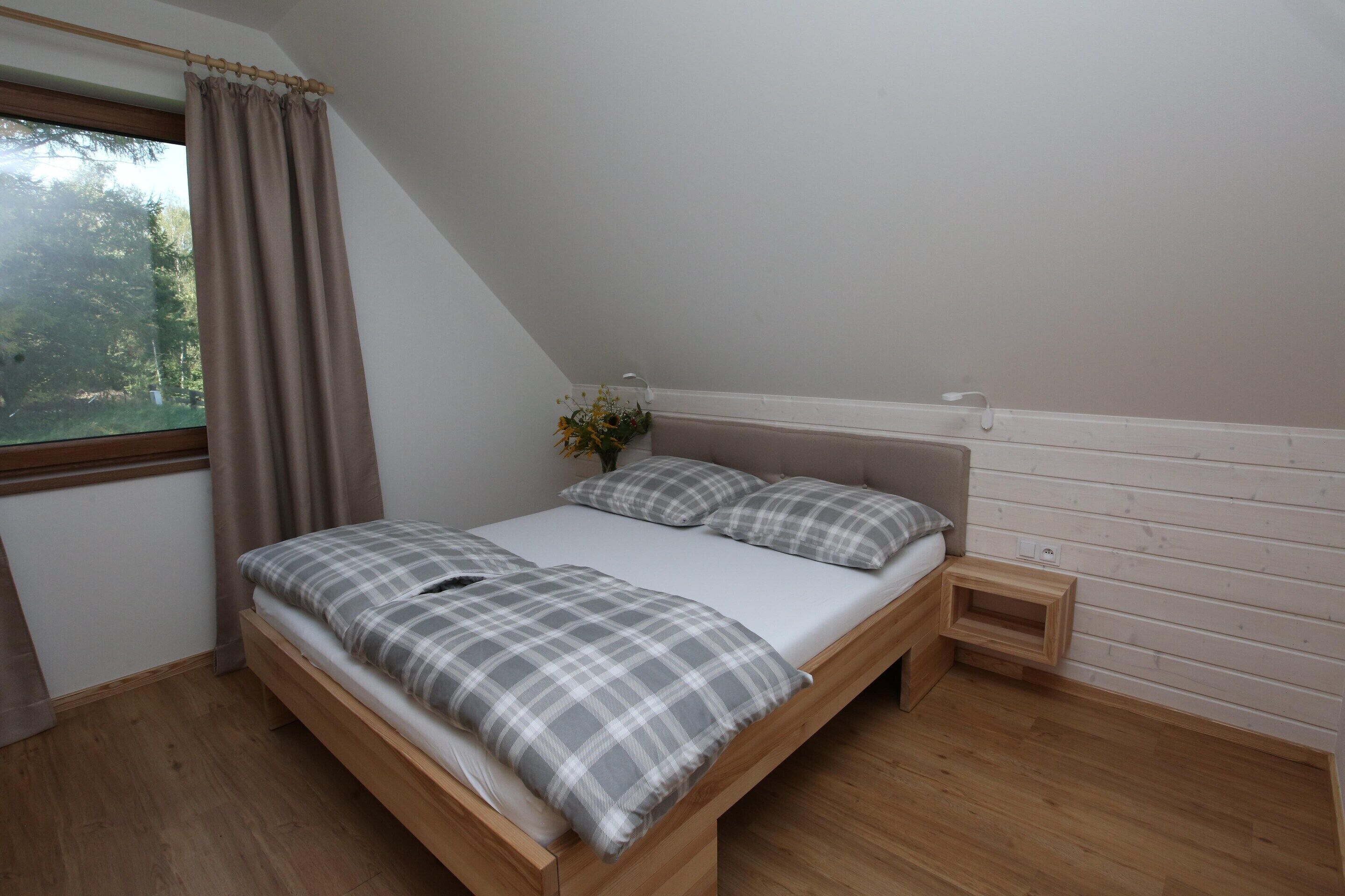 A bedroom with a wooden bed, a window with curtains, a bedside table, and a small wooden shelf.