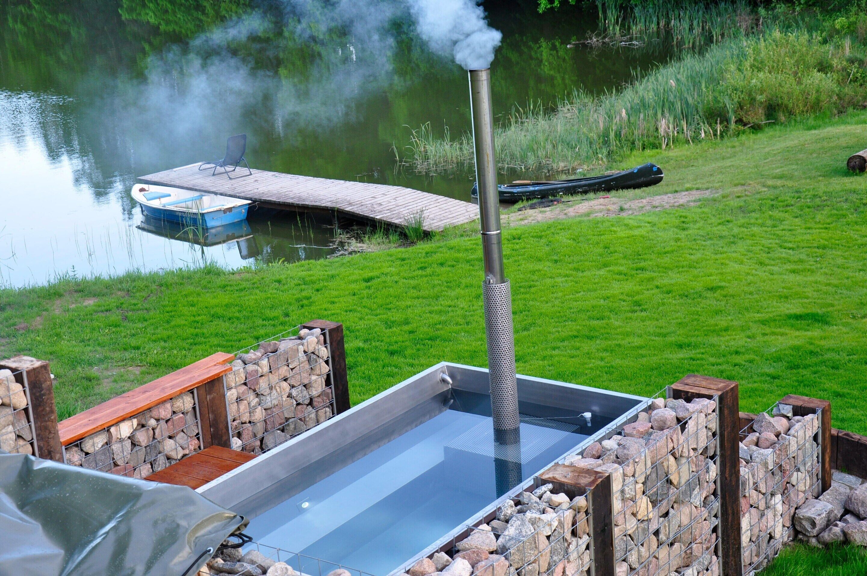 A hot tub with a wooden deck and stone walls, overlooking a calm lake with a dock and a boat.