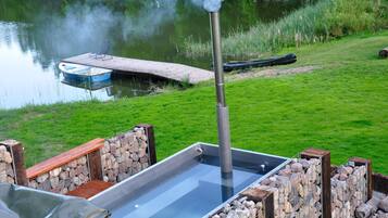 A hot tub with a wooden deck and stone walls, overlooking a calm lake with a dock and a boat.