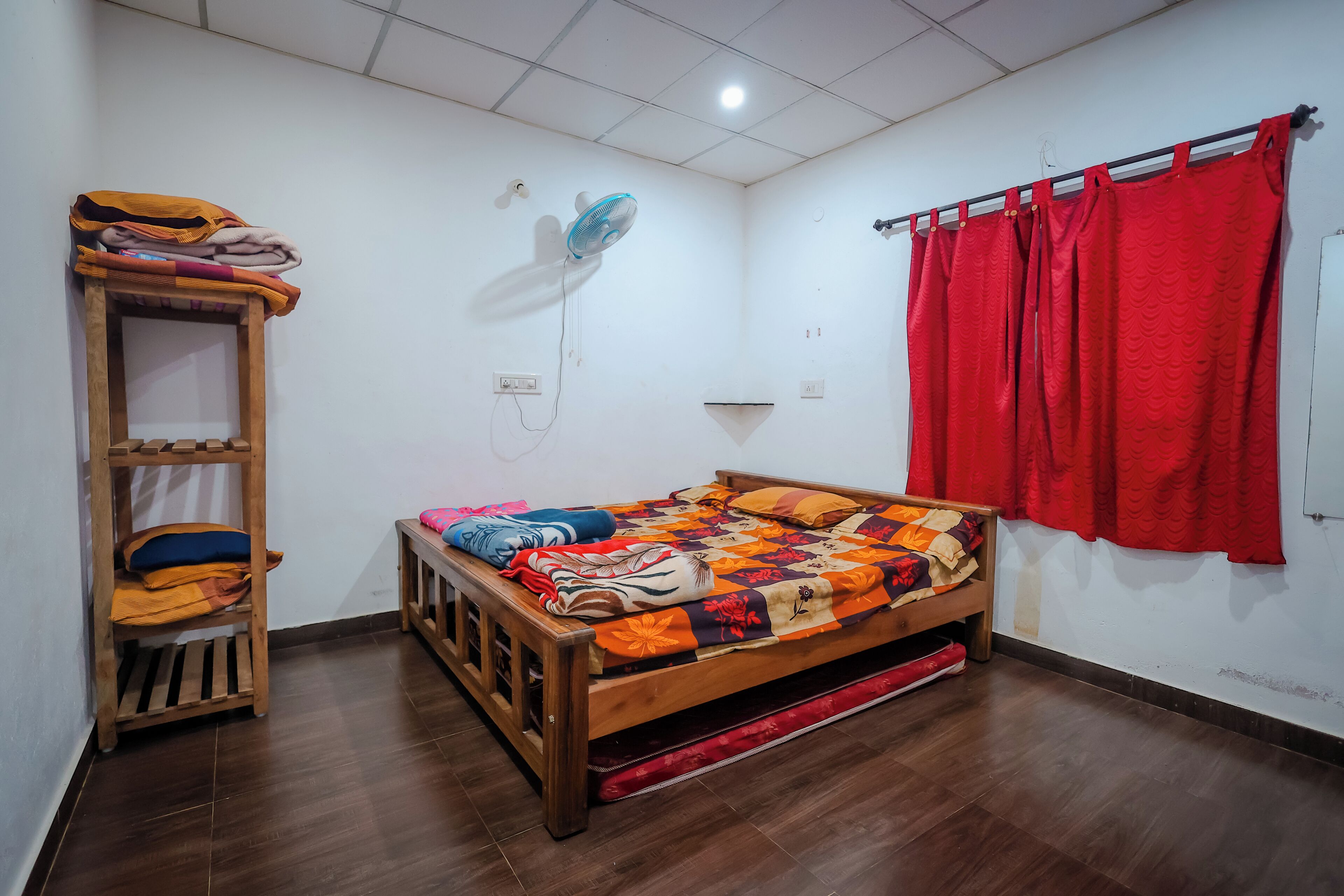 A room with a wooden bed, a wooden shelf, a wall-mounted fan, and a red curtain.