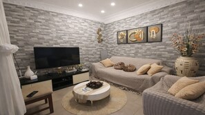 A living room with a brick-patterned wall, a flat-screen TV, a sofa, a coffee table, and a decorative vase with flowers. - Adorable annex for a quiet stay (Conakry)