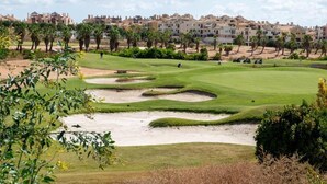 Een golfbaan met zandbunkers, een waterhindernis en op de achtergrond woongebouwen.