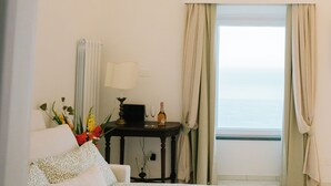 Une chambre d’hôtel avec un lit, une petite table avec une lampe, une bouteille et des verres, un vase avec des fleurs et une vue sur la mer par la fenêtre.