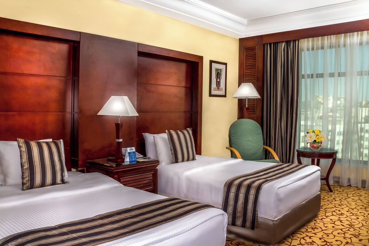 A hotel room with two beds, wooden headboards, a green chair, a small table with a vase of flowers, and a window with curtains.