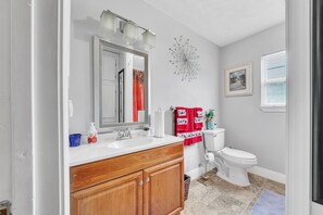 A bathroom with a white sink, wooden vanity, a large mirror, a clock, a toilet, and a window with blinds. - Walk to Beach & Old Town - Bay St. Louis Retreat (Bay St Louis)