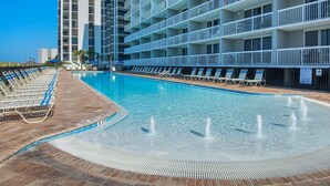 A swimming pool with lounge chairs, a water fountain, and a multi-story building with balconies.