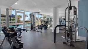 A gym with various exercise equipment, a pool, and large windows offering a view of palm trees.
