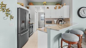 A modern kitchen with a stainless steel refrigerator, a built-in oven, and a toaster. There are two bar stools and a clock on the wall. - 401 Pet-friendly, Pirate's Cove Retreat 3 Bedroom Home (Fort Myers Beach)