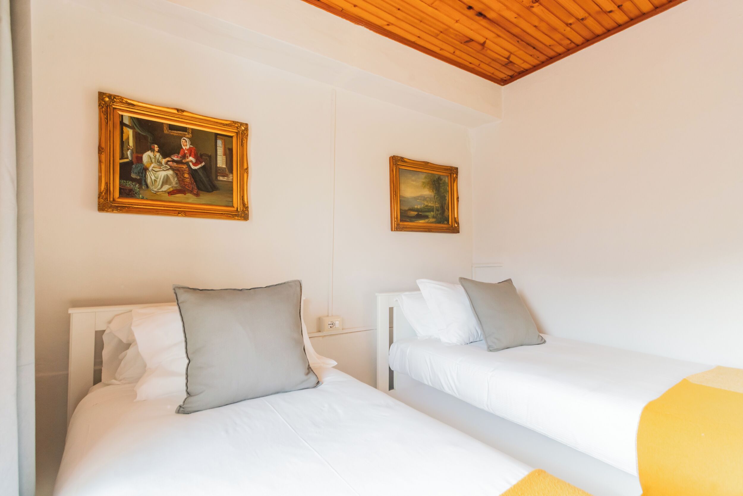 A hotel room with two beds, a wooden ceiling, and two framed paintings on the wall.