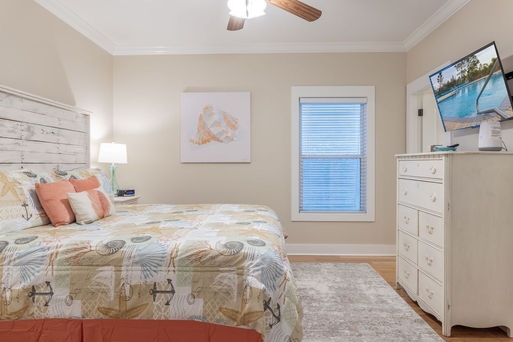 Une chambre à coucher comprenant un lit, une commode, un ventilateur de plafond, une fenêtre avec des stores et un téléviseur fixé au mur.