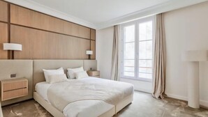 Una habitación de hotel moderna con una cama grande, dos mesitas de noche, un cabecero de madera y un amplio ventanal con cortinas.