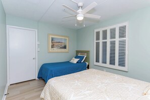 Een slaapkamer met twee bedden, een plafondventilator, een raam met luiken en een lijst met een afbeelding aan de muur.
