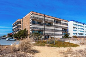 Ett hotell med flera våningar och balkonger, beläget nära en strand med sandig mark och gles vegetation.