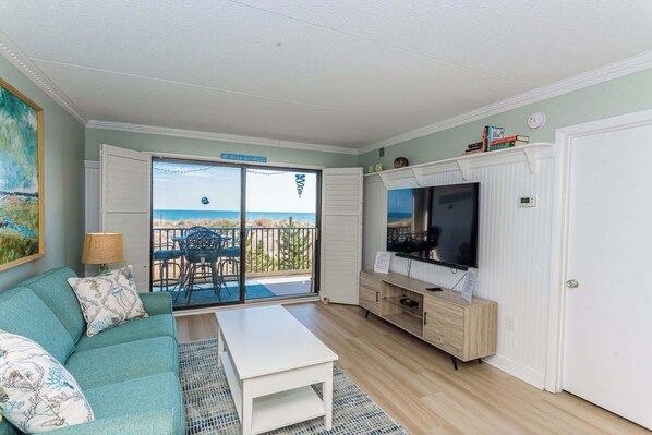 A living room with a sofa, coffee table, and TV stand. There is a sliding glass door leading to a balcony with a view of the ocean.