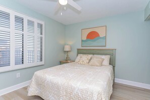 A bedroom with a bed, a ceiling fan, a nightstand with a lamp, and a wall painting.