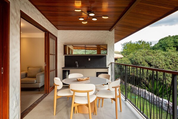 Un espace repas extérieur moderne, doté d’une table ronde et de six chaises, d’un barbecue intégré et offrant une vue sur un jardin luxuriant.