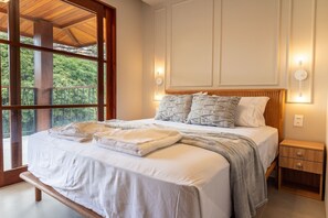 Un dormitorio con una cama grande, cabecera de madera, mesitas de noche y vistas a la vegetación a través de amplios ventanales.