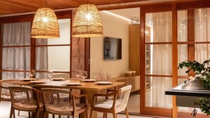 A dining area with a wooden table and chairs, two woven pendant lights, and a view of the kitchen through a glass door.