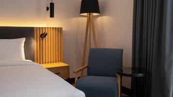 A hotel room with a bed, a chair, a nightstand, a lamp, and a small table.