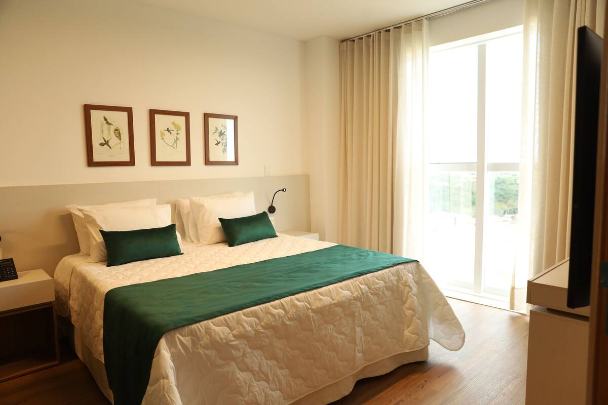A neatly made bed with a green and white bedspread, a bedside table with a phone, and a window with curtains.