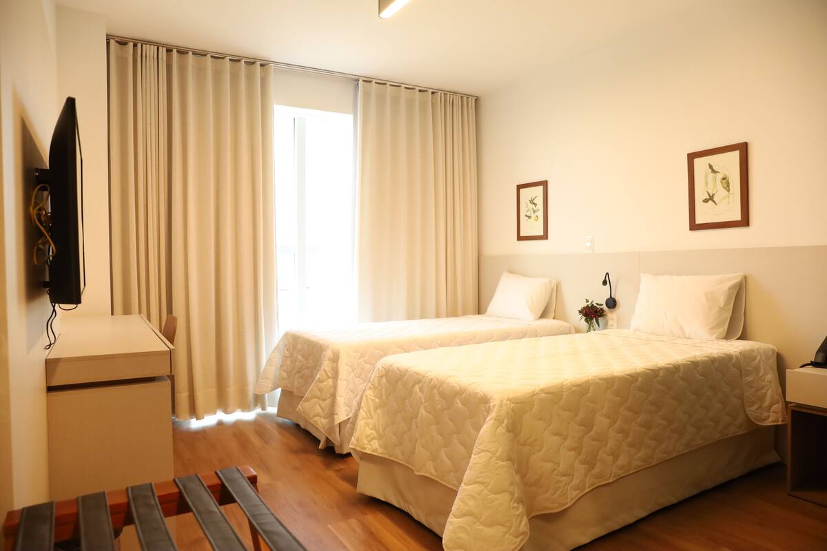 A hotel room with two beds, a desk, a TV, and a window with curtains.
