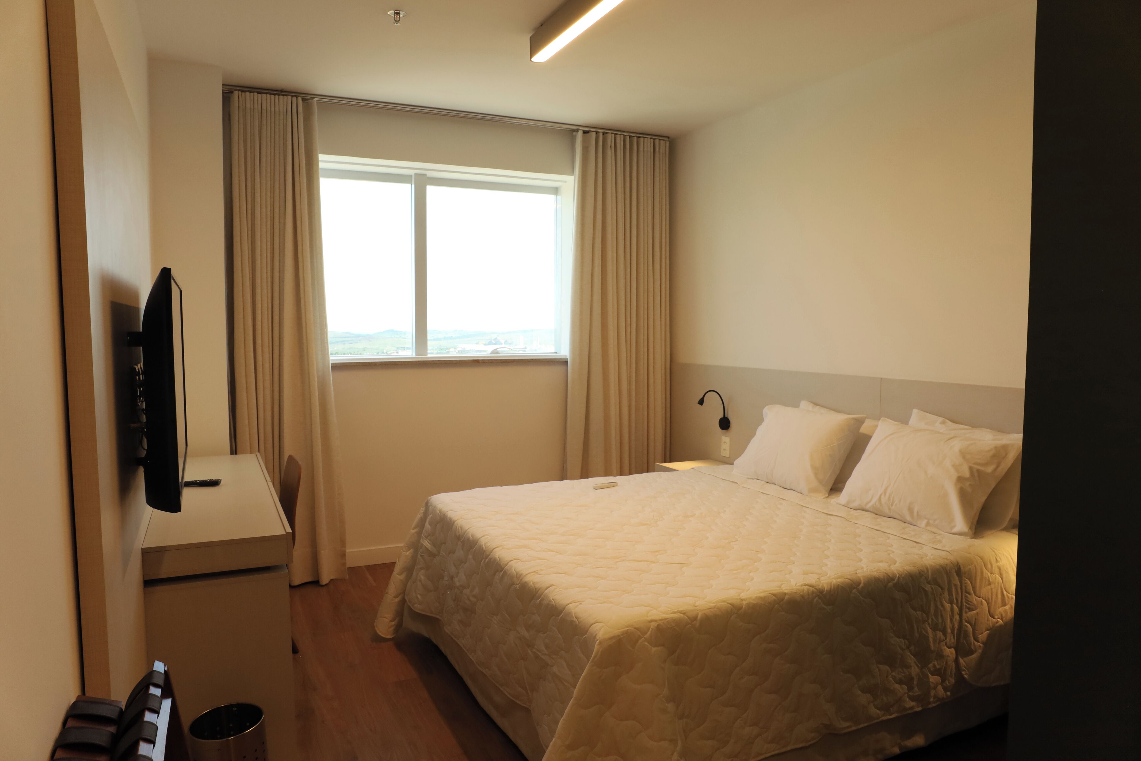 A hotel room with a bed, a desk with a television, a window with curtains, and a nightstand with a lamp.