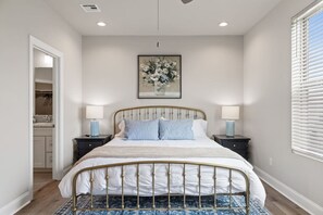 Un dormitorio con una cama, dos mesitas de noche, una mesita auxiliar, una obra de arte enmarcada en la pared y una ventana con persianas.