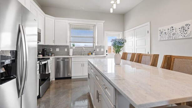A modern kitchen with white cabinetry, stainless steel appliances, a marble countertop, and a dining area with wicker chairs.