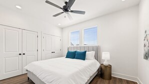 Une chambre à coucher avec un lit, des murs blancs, un ventilateur de plafond et une fenêtre avec des stores.
