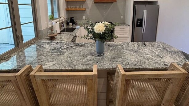 A kitchen with a granite island, wooden chairs, pendant lights, a refrigerator, and a window with blinds.