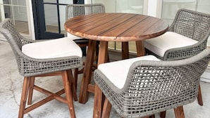 A round wooden table with four wicker chairs, each with a white cushion. The setting is outdoors with a concrete floor and a glass door in the background.
