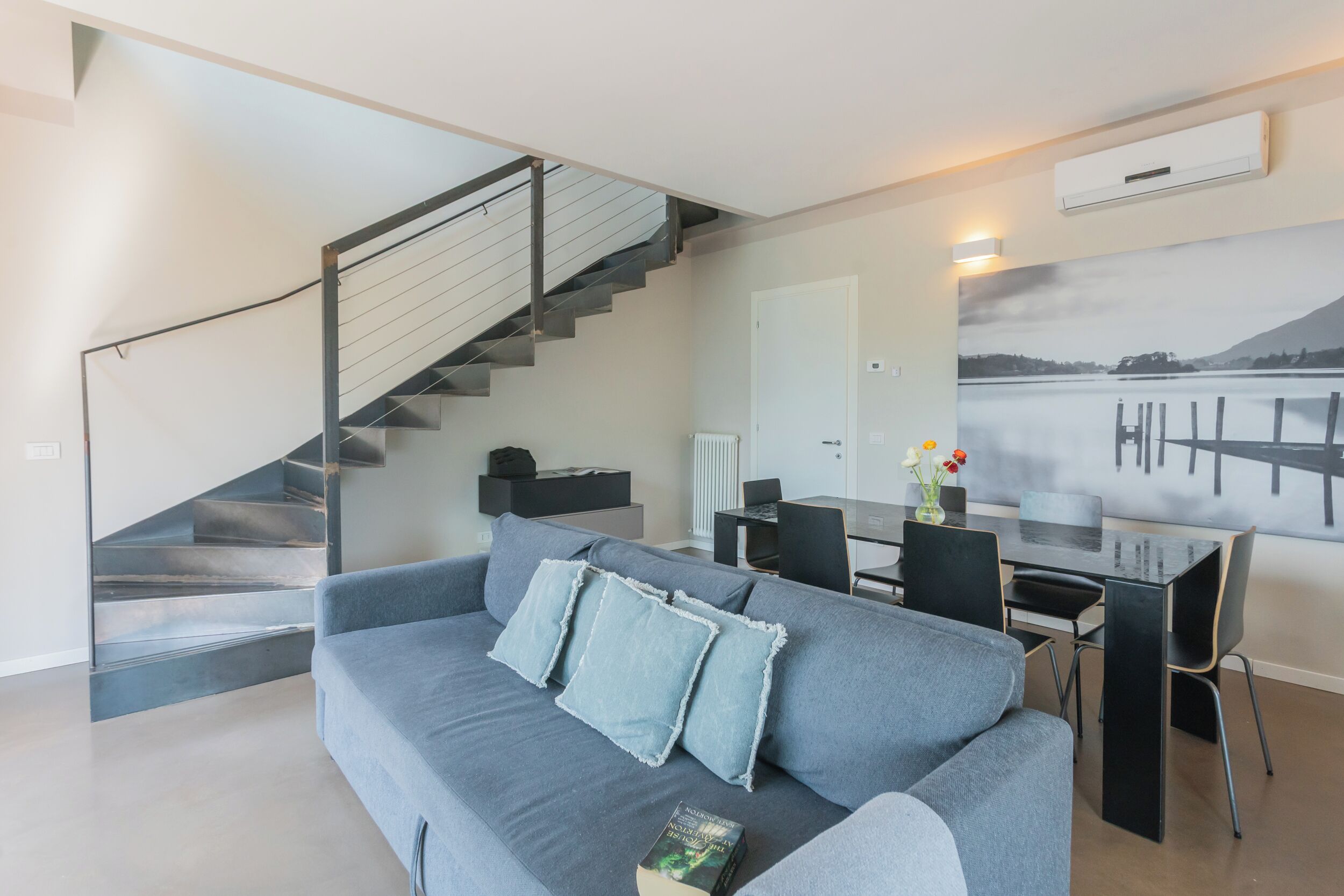 Een moderne woonkamer met een grijze bank, een trap, een eethoek en een groot landschap op de muur.