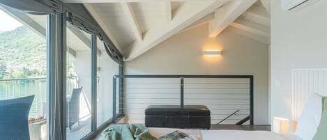 A modern bedroom with a large window, a bed, a reading area, and a balcony with outdoor furniture.