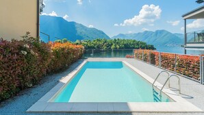 A rectangular swimming pool with a clear blue water surface, surrounded by a metal railing and a hedge with autumn-colored foliage. In the background, there are mountains and a body of water, suggesting a scenic location. - Mulino Vista Lake by Rent all Como (Tremezzina)