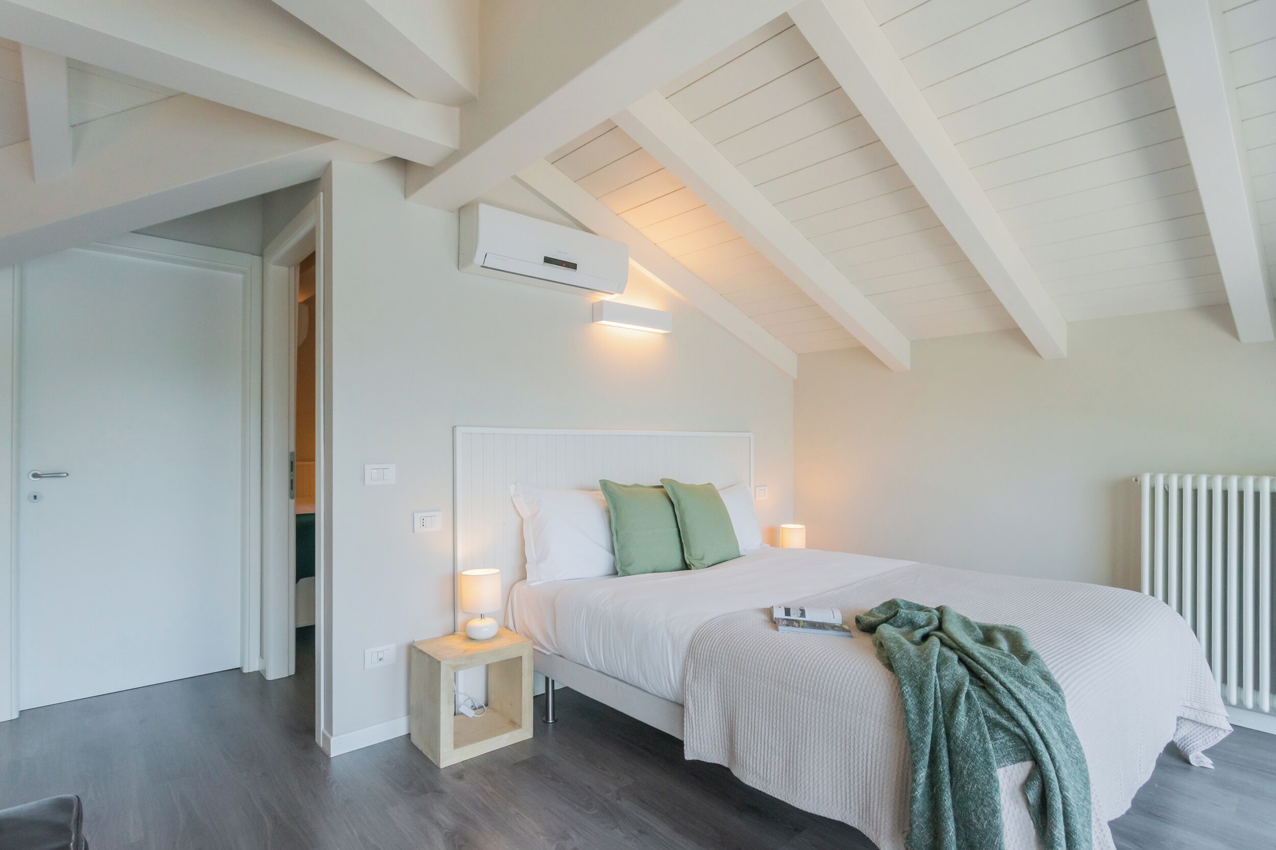 Een slaapkamer met een schuine kap, een bed met wit beddengoed, twee bedlampen, een nachtkastje en een wandairconditioner.
