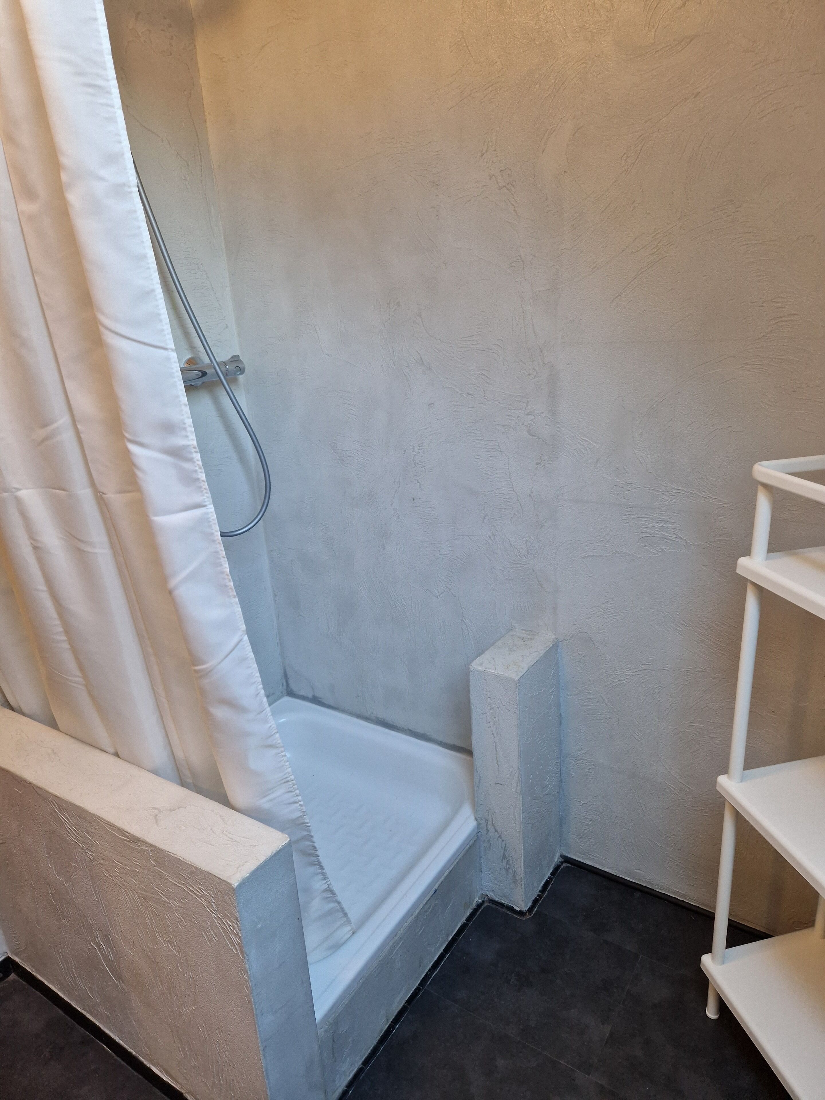 Un petit espace douche fermé, avec un rideau de douche blanc, une étagère blanche et un sol carrelé.