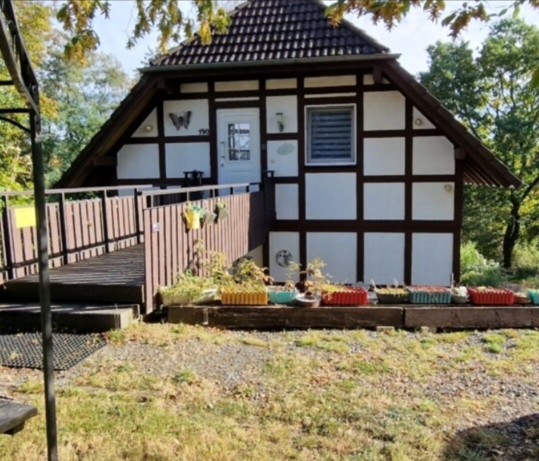 A traditional half-timbered building with a wooden door and windows, surrounded by a garden with various plants and a wooden fence.