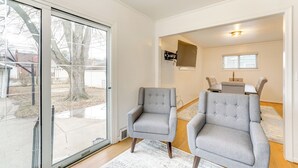 A spacious living room with two grey armchairs, a dining area with a table and chairs, and a sliding glass door leading to a patio. - 13 Mi to Downtown Detroit: Bright Home (Dearborn)